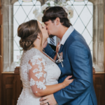 Elopement Wedding A bride and groom share a kiss indoors in front of a decorative window. The bride is wearing a white lace wedding dress with a sheer, floral-embroidered overlay and has her hair styled in an updo. The groom is in a blue suit with a white shirt. Both have their arms wrapped around each other. Elopements Inc